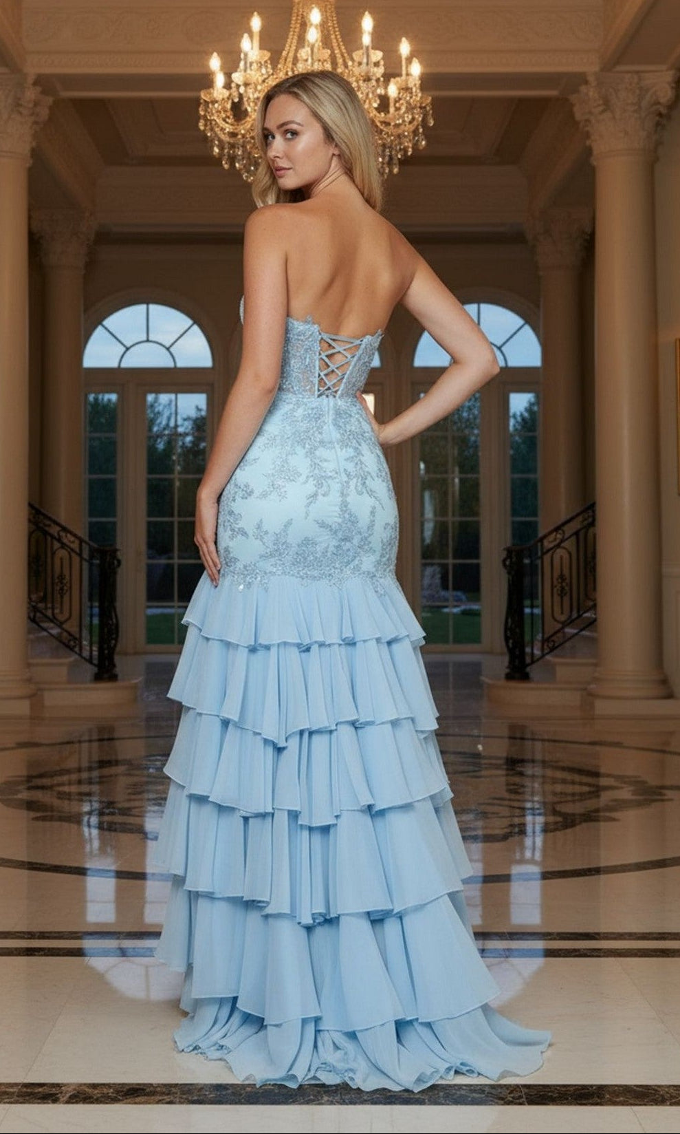A woman in the Long Prom Dress: Poly USA 25066, featuring a strapless lace embroidered corset bodice and tiered ruffled chiffon skirt, stands in an elegant hall with marble floors and a chandelier.